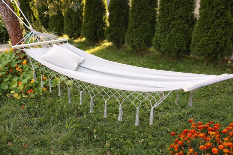 Platnena ljuljaška sa jastukom postavljena između drveća u uređenom dvorištu okruženom travom i cvećem